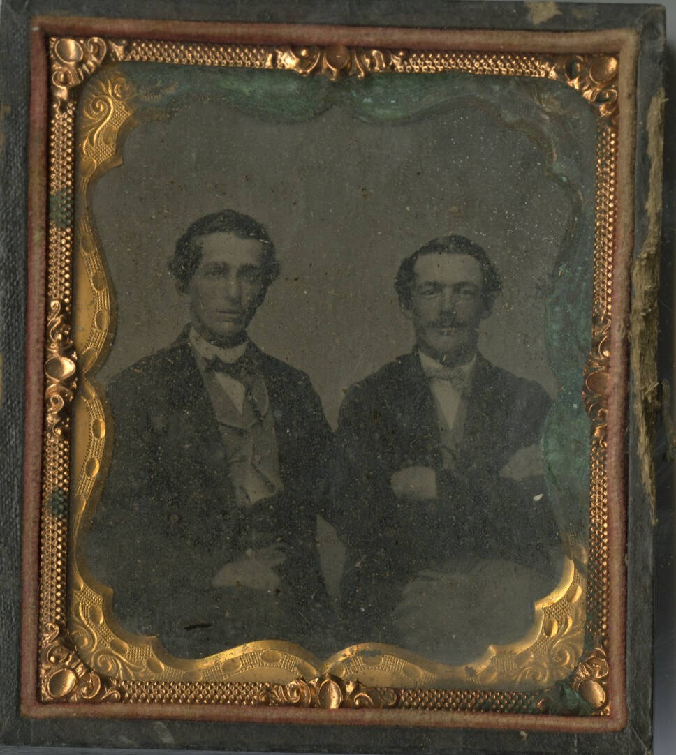 Black and white ambrotype photographic portrait of two seated men, one has his arms crossed.
